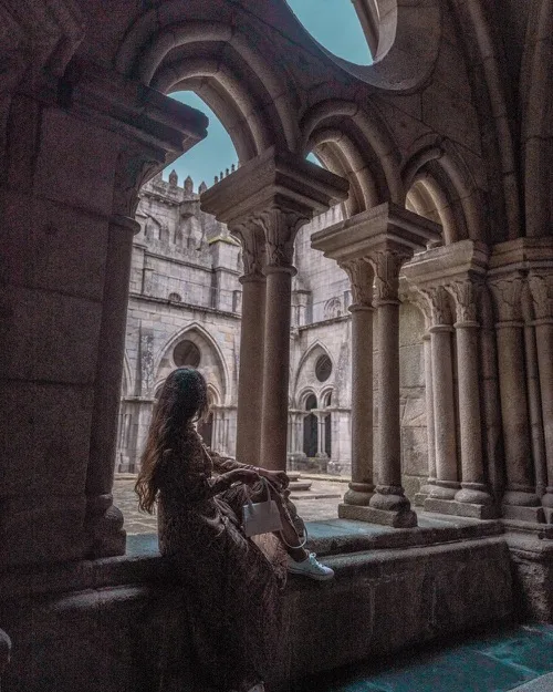 Woman sitting on a cloister ledge, silhouetted against Gothic arches and a stone courtyard in soft daylight