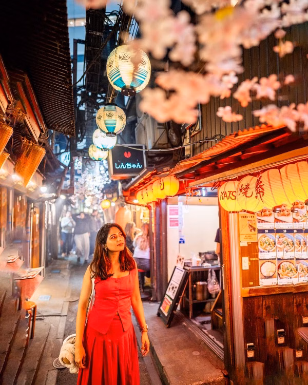 Woman in a red dress stands in a lantern-lit alley lined with restaurants and cherry blossoms
