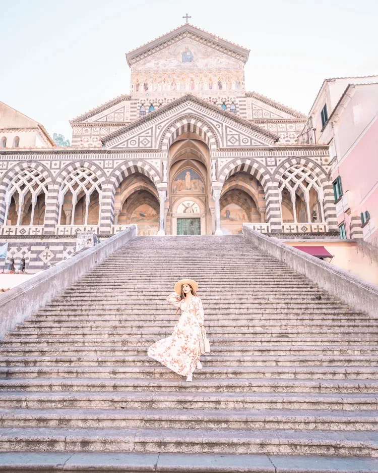 Woman in a flowing floral dress and straw hat descending wide stone steps toward the striped Romanesque façade of Amalfi Cathedral.