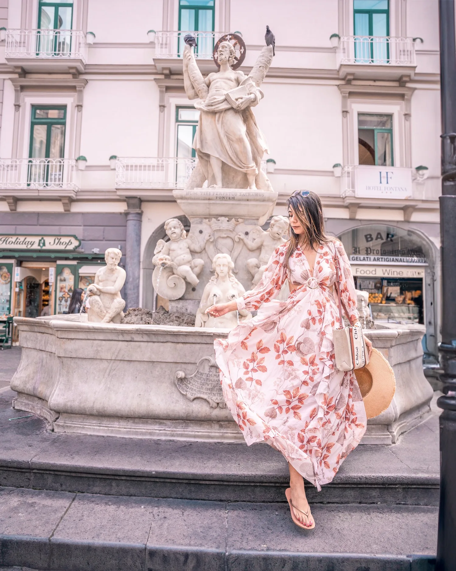 Woman in a flowing floral dress posing by an ornate stone fountain in a European city square, holding a straw hat and tote bag.