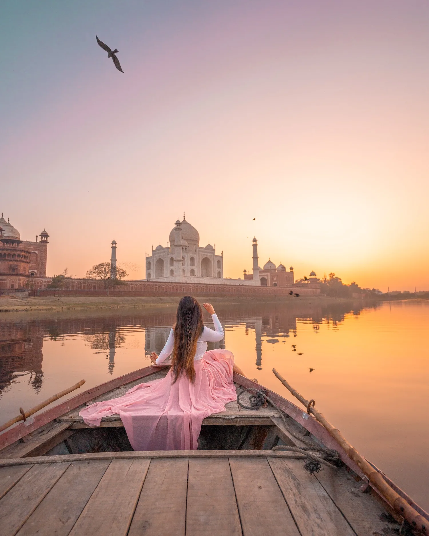 Woman in a flowing pink skirt seated in a wooden boat at sunrise, gazing across the river at the Taj Mahal.