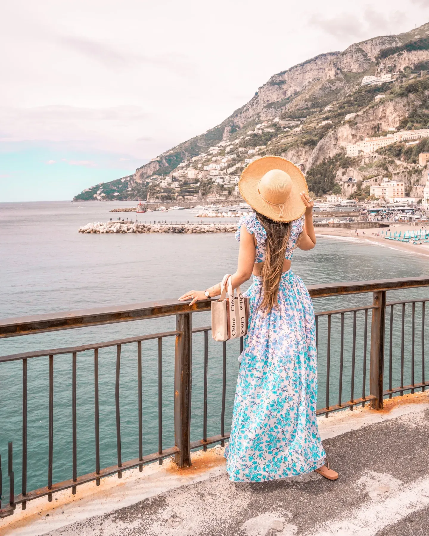 Woman in a floral maxi dress and wide straw hat gazes over a coastal town and turquoise sea from a cliffside railing.