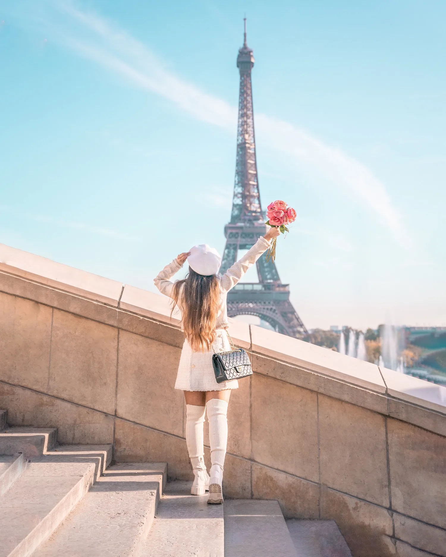 Woman in a white beret and coat holds a bouquet toward the Eiffel Tower from stone steps on a sunny day.