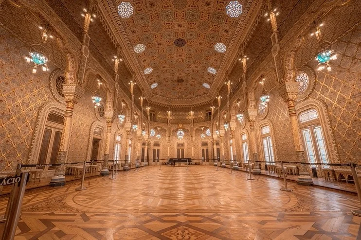 Lavishly gilded, high-ceilinged ballroom with intricate patterned walls, ornate columns and a polished parquet floor bathed in warm light