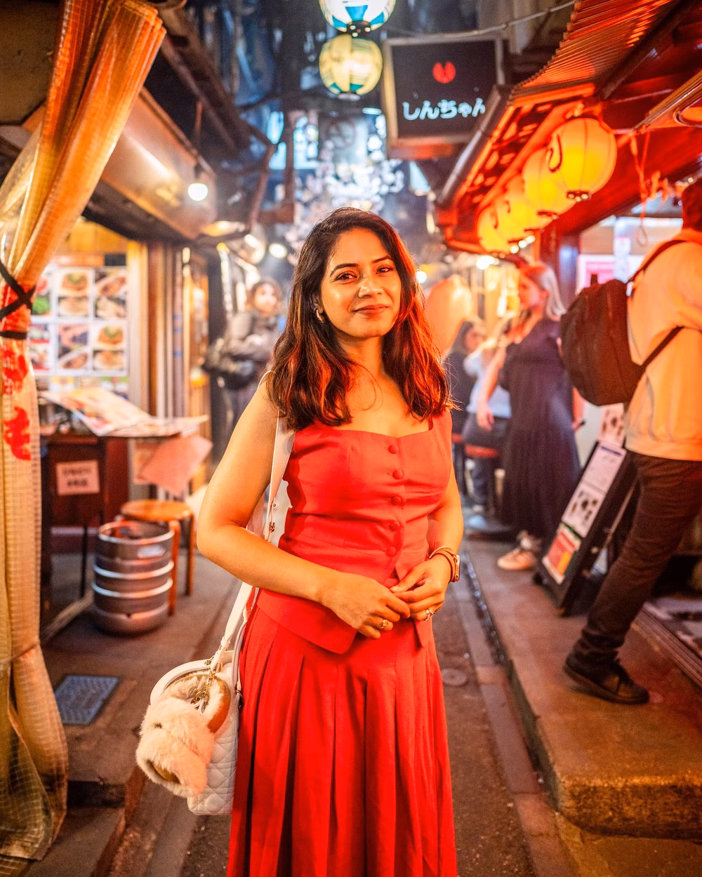 A woman in a red dress stands in a lantern-lit alley market at night