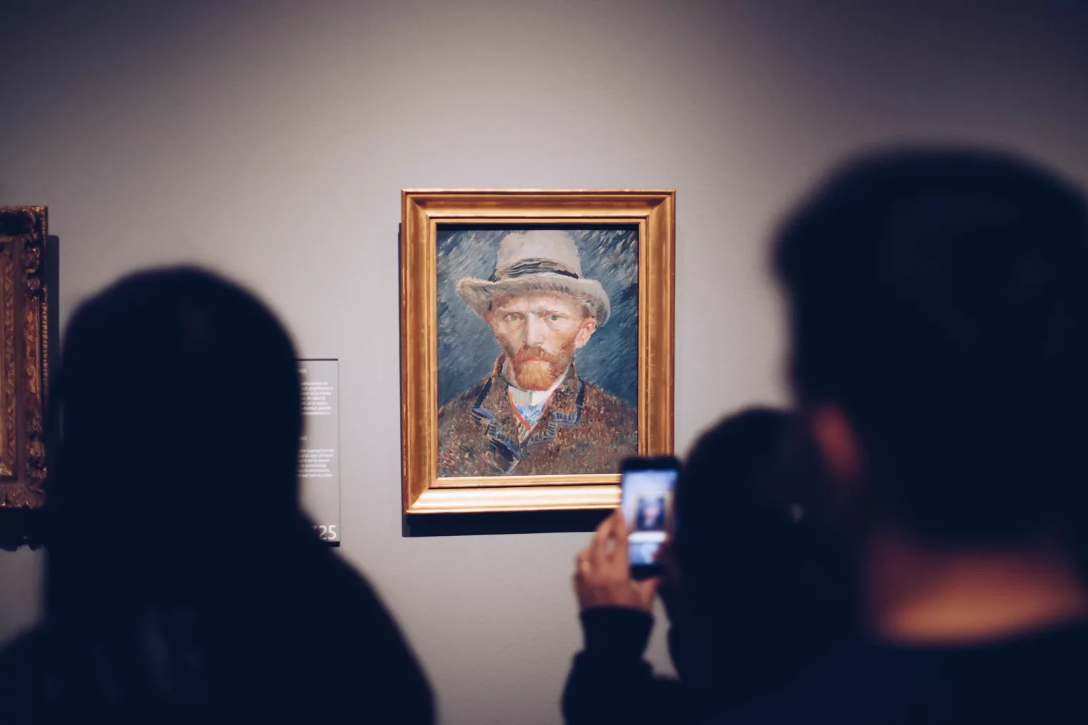 Visitors photographing a framed self-portrait of a bearded man wearing a hat displayed on a museum wall