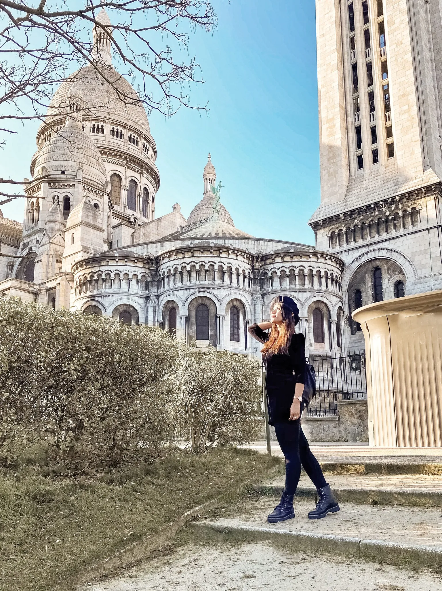 Woman in black outfit and beret posing by the Sacré-Cœur Basilica with its domes and towers under a clear blue sky