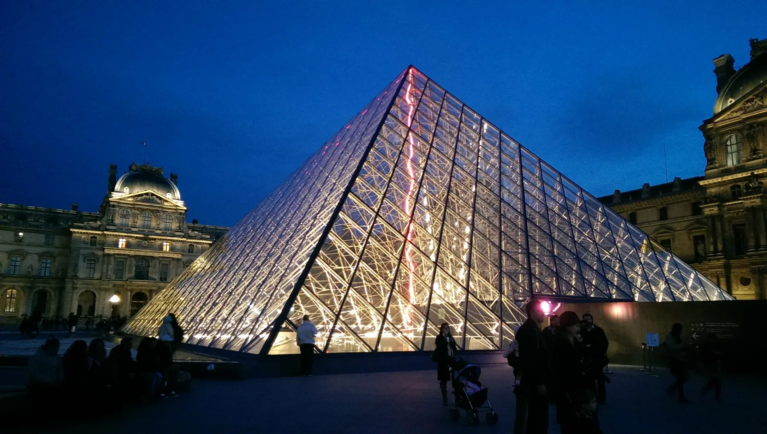 Glass pyramid entrance to the Louvre lit from within against a deep blue evening sky with surrounding historic museum façades.