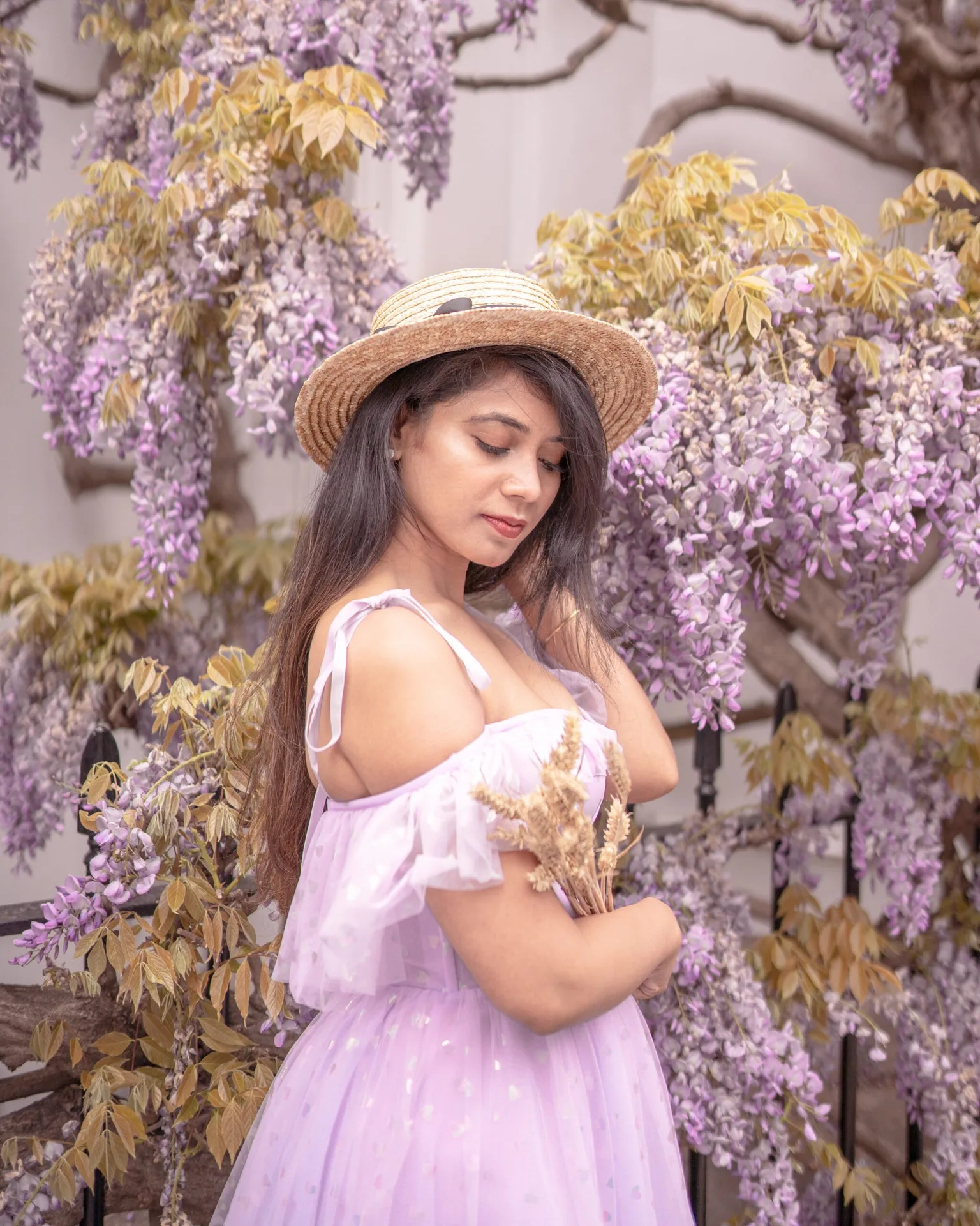 Young woman in a straw hat and lilac off-shoulder dress holding dried flowers beside cascading wisteria blooms.