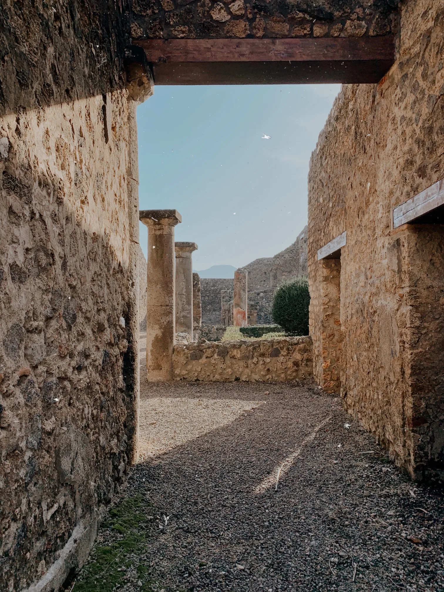 Sunlit ancient stone ruins framed by a narrow passageway leading to weathered columns and distant hills.