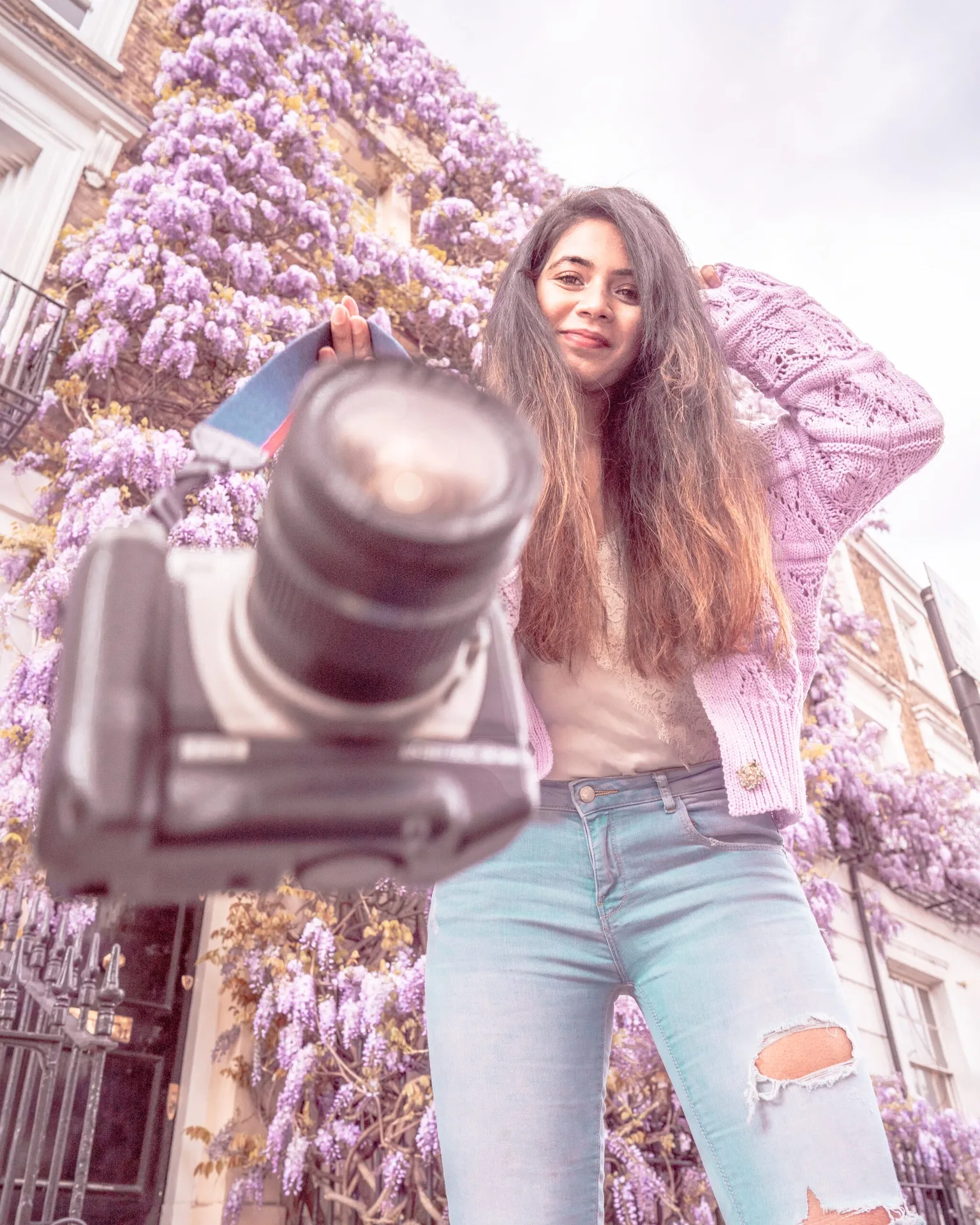 Woman in a lavender cardigan smiling toward the camera while handing a DSLR in front of a row of wisteria-covered townhouses.