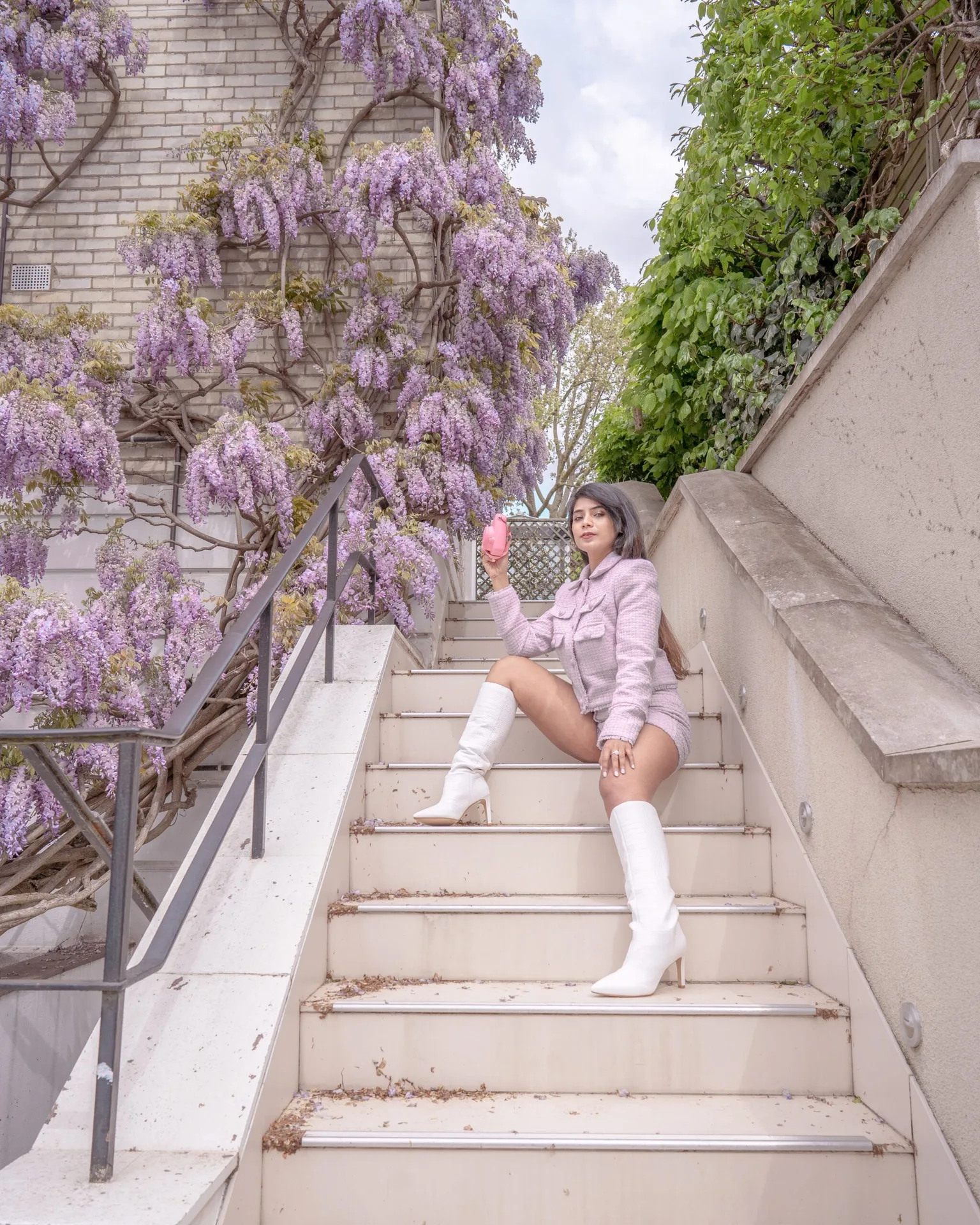 Woman in a pink tweed jacket and white knee-high boots sitting on outdoor steps beside cascading purple wisteria, holding a small pink camera.