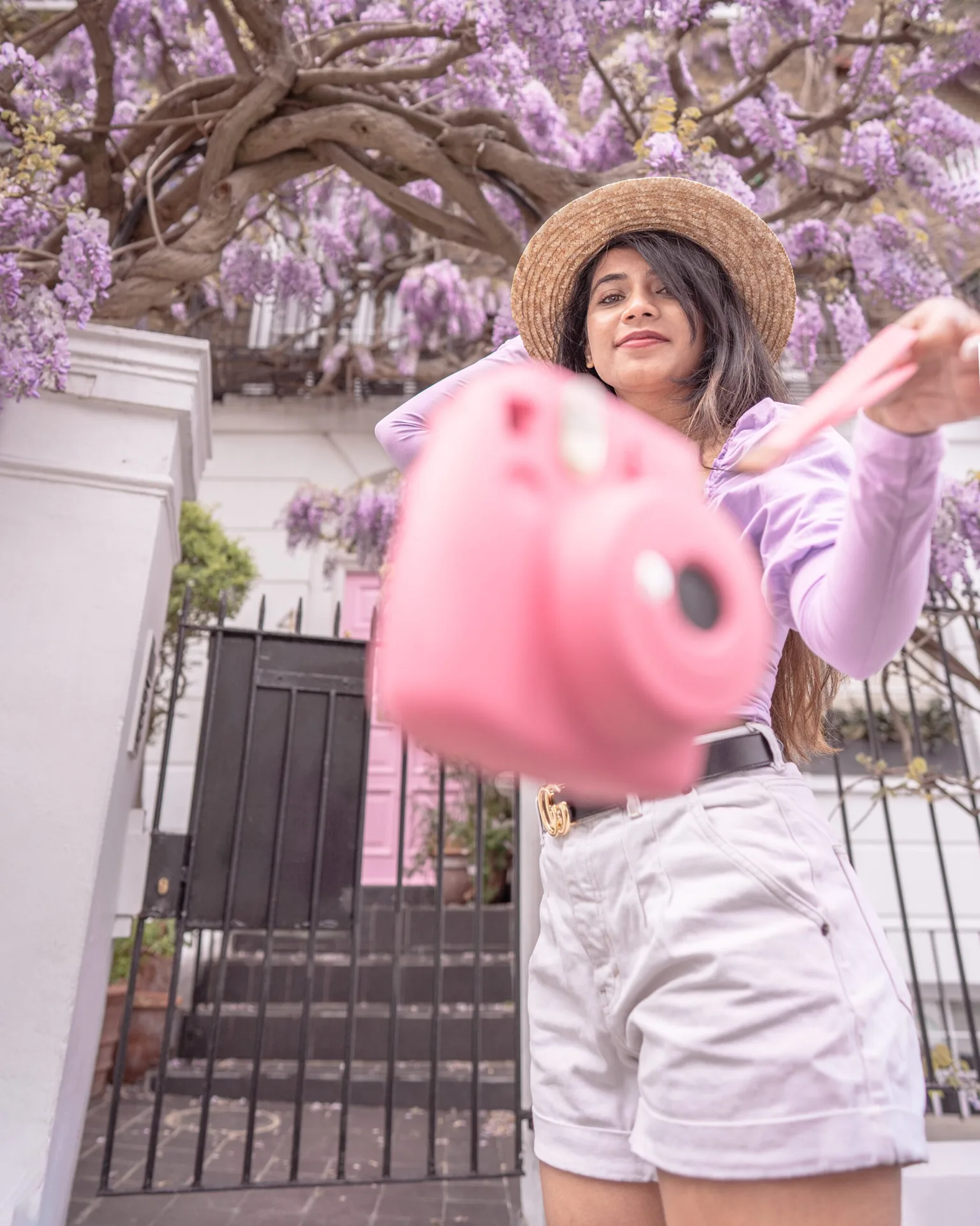 Woman in a straw hat holding a pink instant camera toward the viewer beneath hanging purple wisteria on a pastel-fronted street