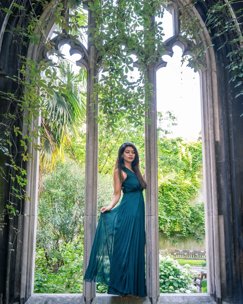 Woman in a flowing teal dress posing gracefully in a tall, ivy-framed Gothic window overlooking a lush garden.