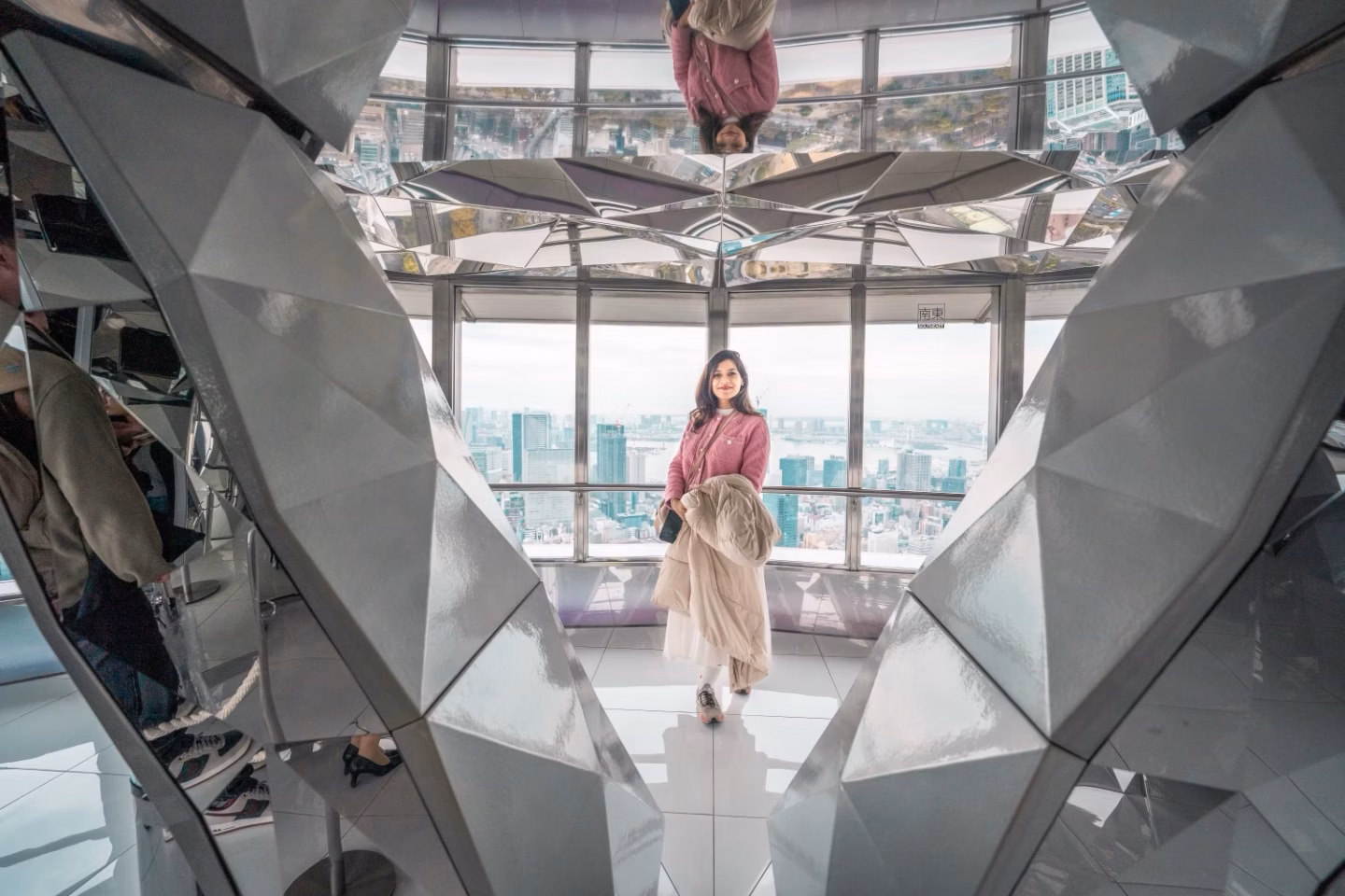 A woman stands in a mirrored observation deck with a city skyline visible through the windows