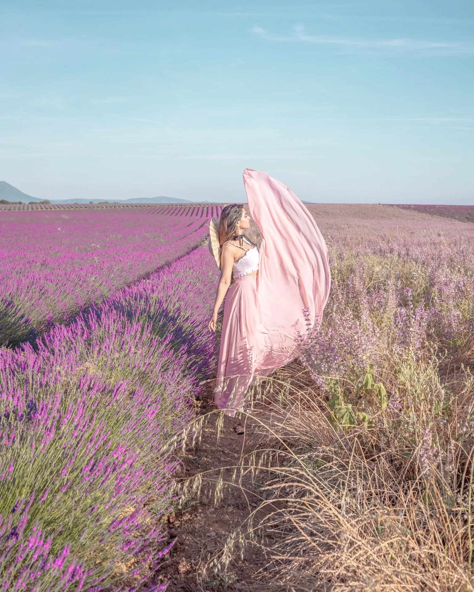 Woman in a flowing pink dress standing in a vast lavender field with rows of purple blooms stretching to the horizon.