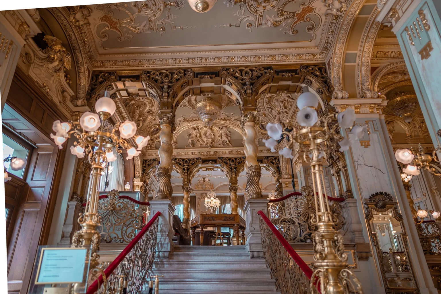 Lavish gilded staircase and balustrades leading to an ornate, chandelier-lit salon with carved columns and decorative plasterwork.