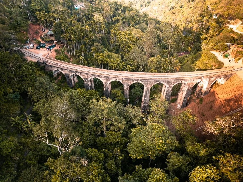 Curved stone railway viaduct spanning a lush green valley with tall trees and sunlit hillsides.