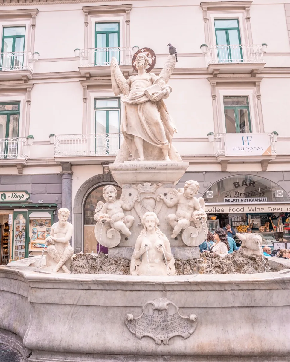 Marble fountain with a central statue of a robed figure surrounded by cherubs and water spouts in a sunny town square.