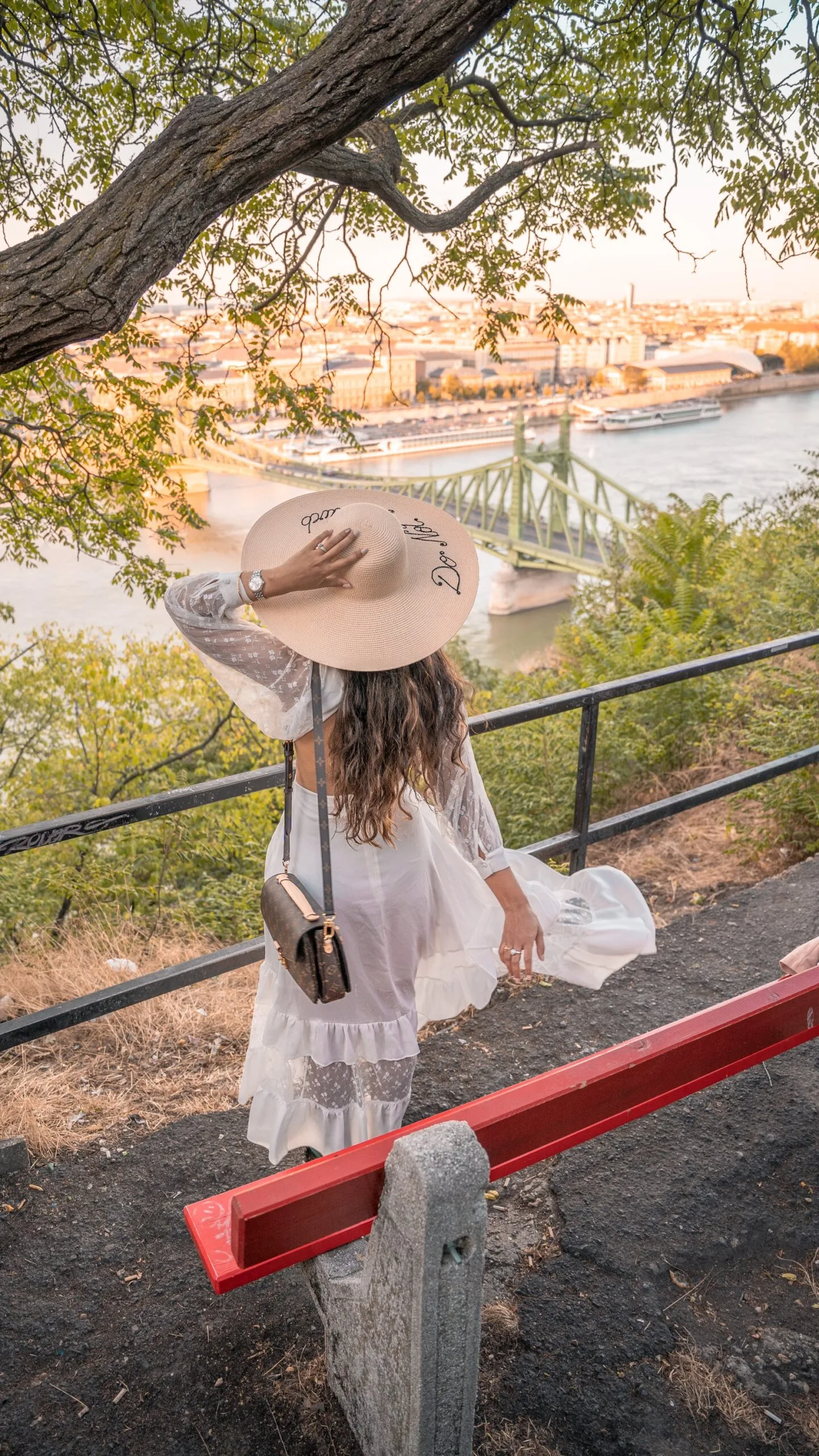Woman in a white dress and wide-brimmed hat overlooking a river and green suspension bridge from a leafy hillside viewpoint.