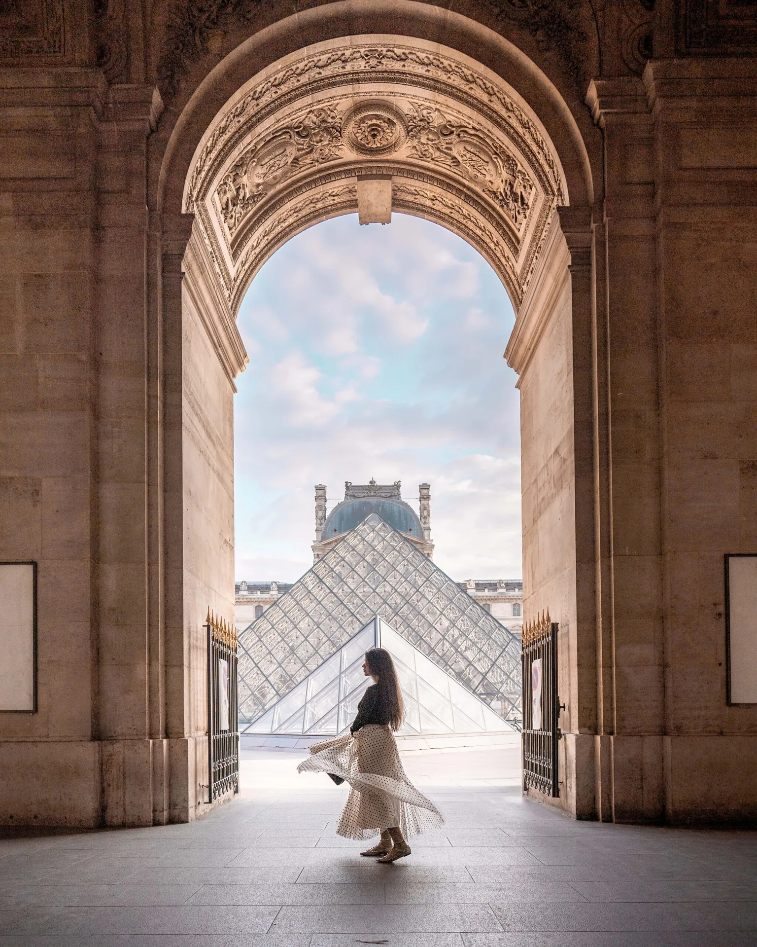 Woman twirling in a flowing skirt beneath a grand archway with the Louvre glass pyramid framed in the background.