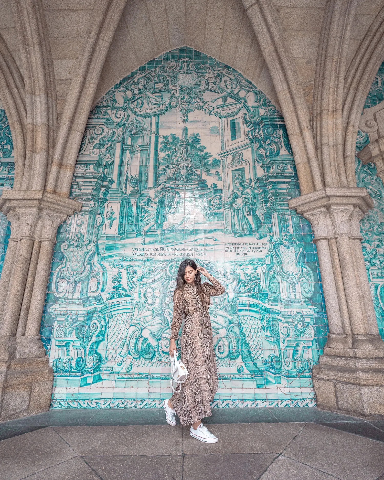 Woman in a patterned maxi dress and white trainers posing under ornate turquoise azulejo tile panels framed by stone arches.