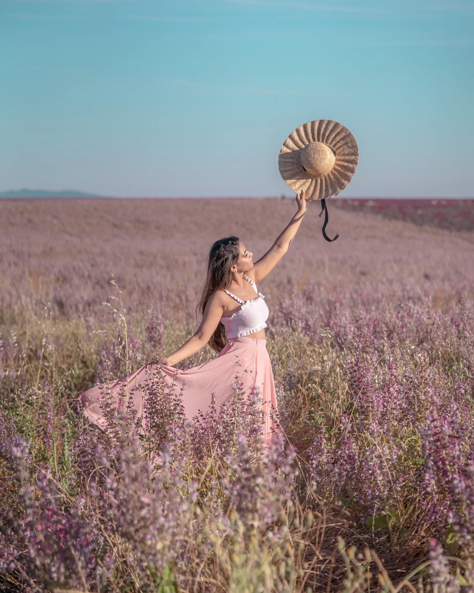 Woman in a flowing pink skirt and white crop top holding a wide straw hat aloft while standing in a lavender-coloured wildflower field under a clear blue sky