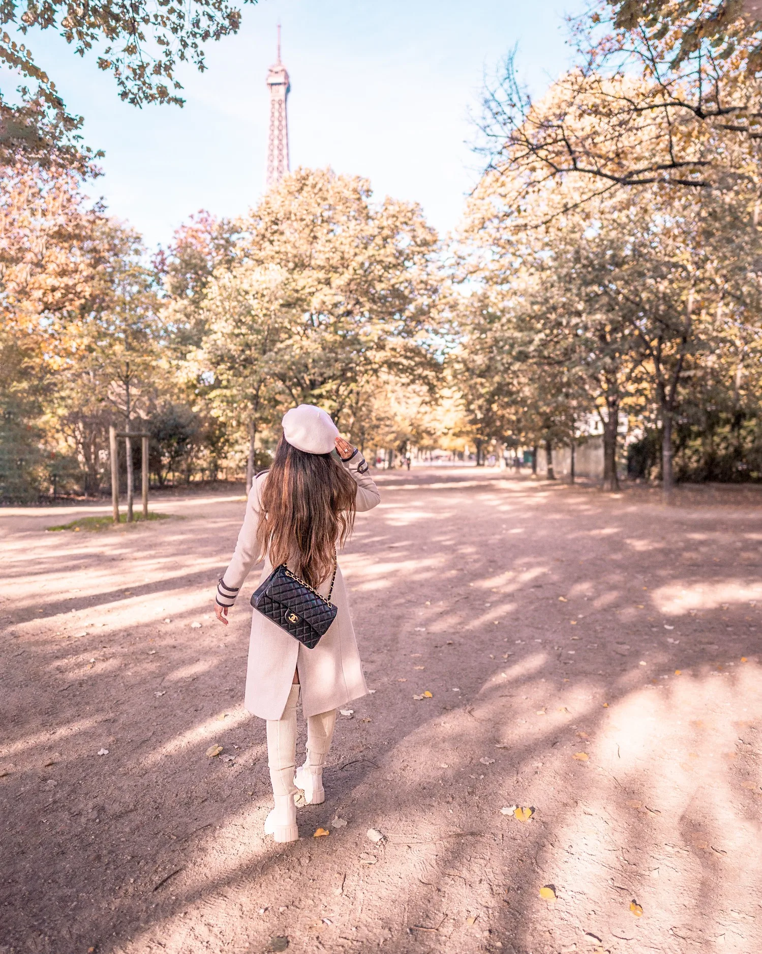 Woman in a beige coat and beret walking down a tree-lined Paris path with the Eiffel Tower visible above the autumn foliage