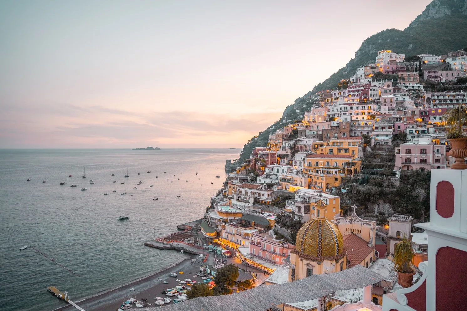 Sunset view of pastel cliffside houses and the tiled church dome of Positano overlooking calm boats on the Amalfi Coast.