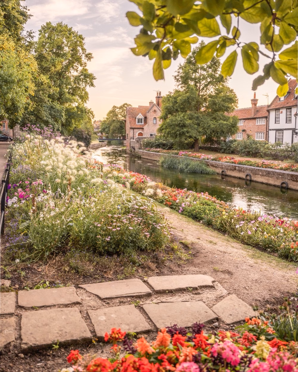 A flower-lined canal runs past charming brick houses and leafy trees in a peaceful village setting