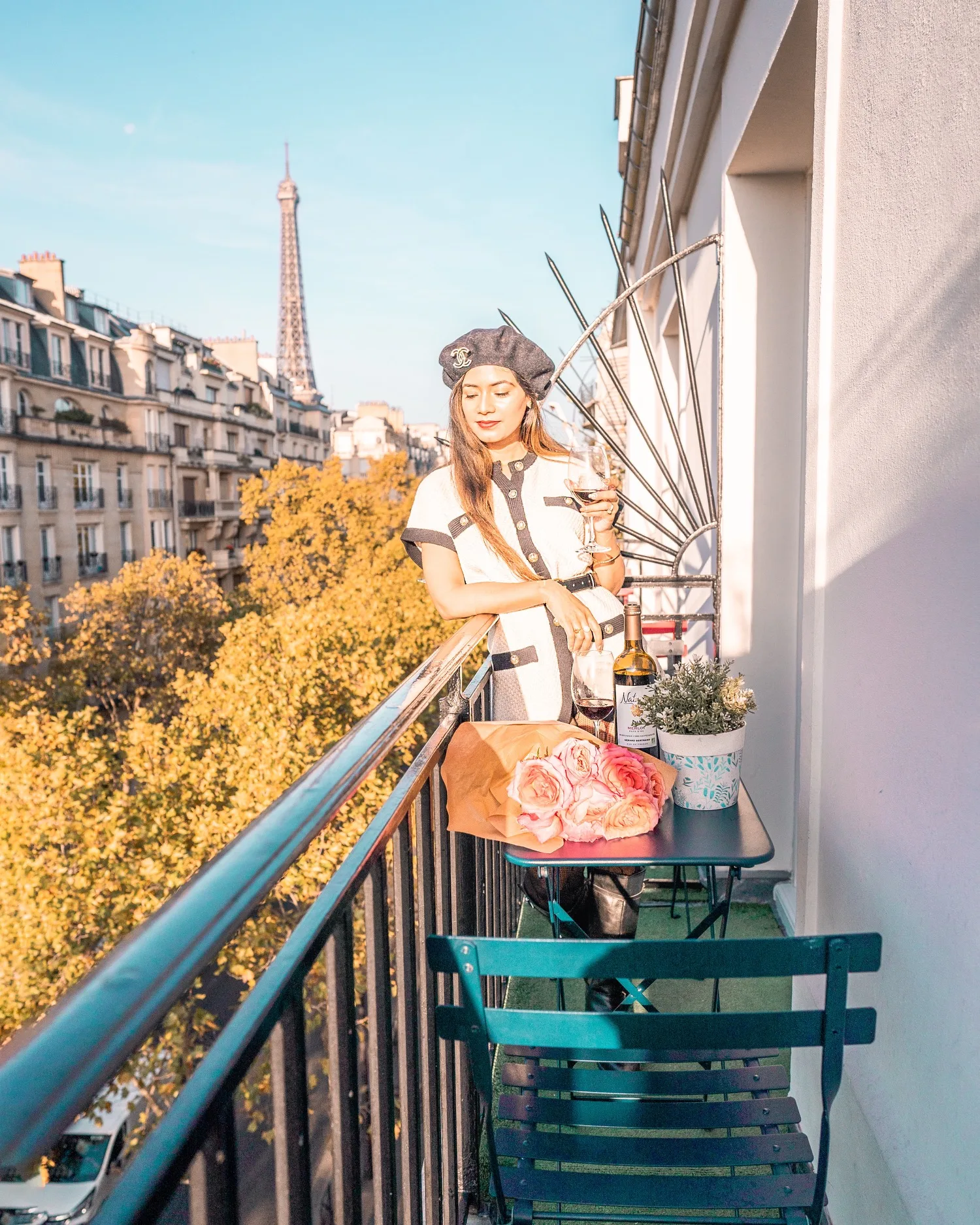 Woman in a beret sipping wine on a narrow Paris balcony with a table of roses and the Eiffel Tower visible in the background