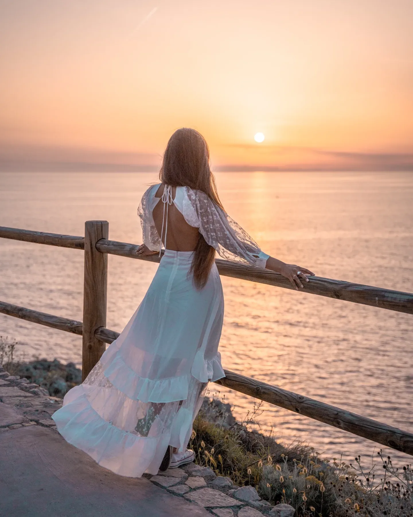 Woman in a flowing white dress leaning on a wooden railing and watching the sun set over a calm sea.