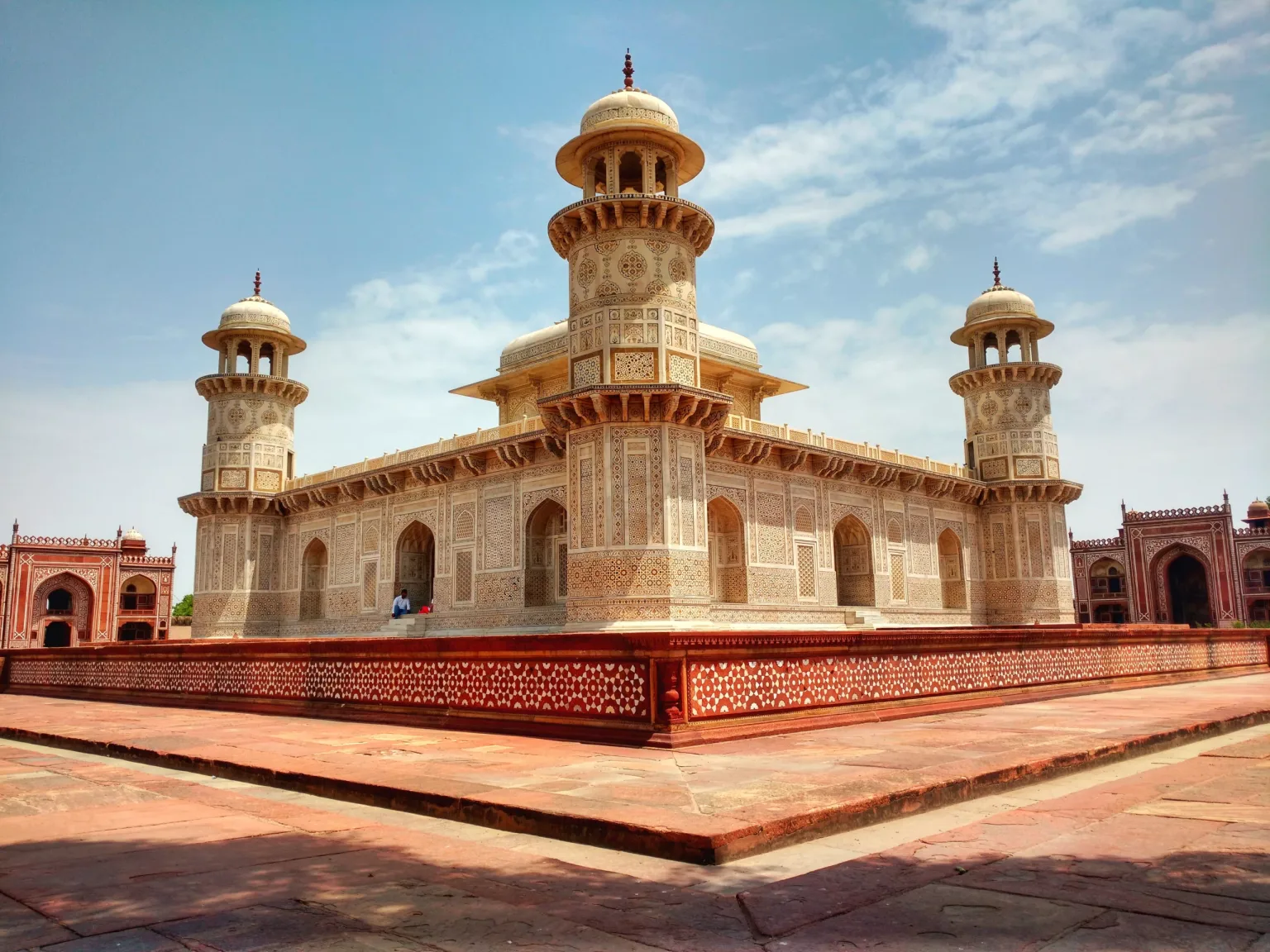 Ornately carved white marble mausoleum with four domed corner towers set on a red sandstone platform under a blue sky.