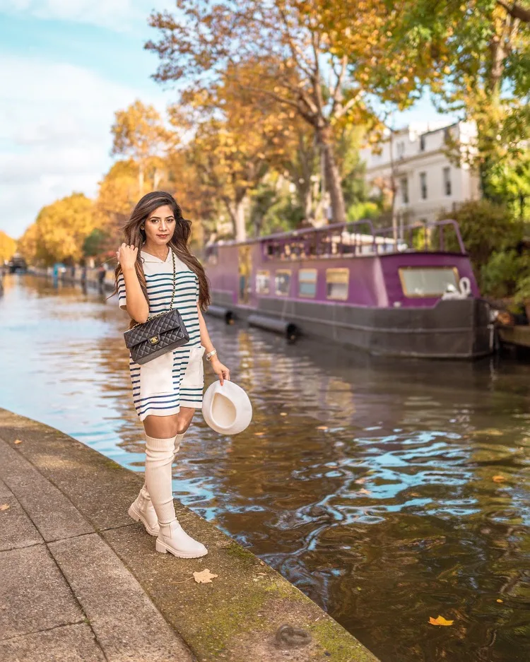Woman in a striped dress and knee-high boots holding a white hat while standing beside a canal with a purple houseboat and autumn trees in the background.
