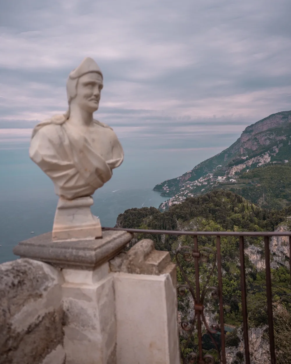 Marble bust on a terrace railing overlooking a dramatic coastal cliff and turquoise sea under a cloudy sky.