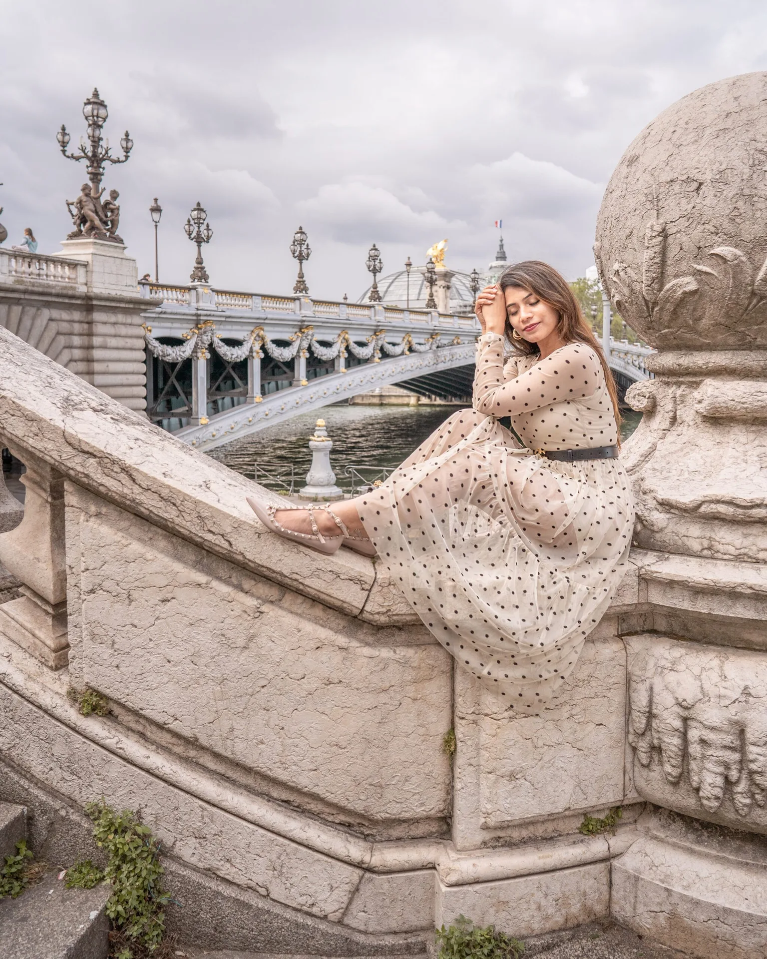 Woman in a polka-dot dress sitting on ornate stone steps beside a river with a decorative bridge in the background.