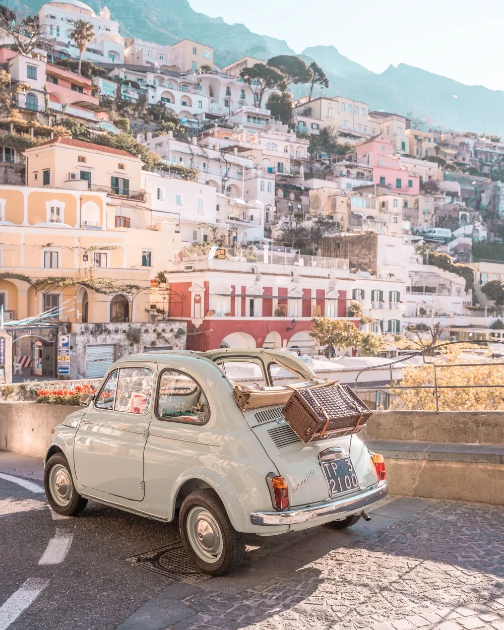 Vintage pale blue Fiat 500 with a wicker picnic basket on its rear rack parked on a coastal road overlooking pastel cliffside houses.