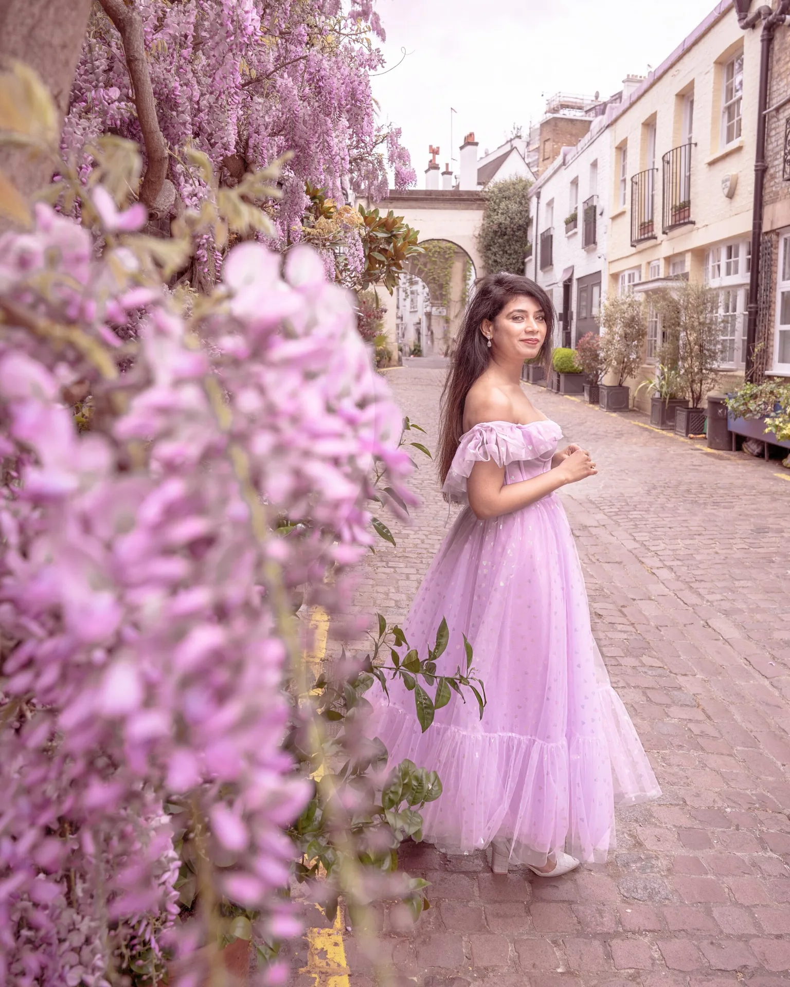 Woman in a flowing lavender gown standing on a cobbled mews lined with pink wisteria and pastel townhouses.