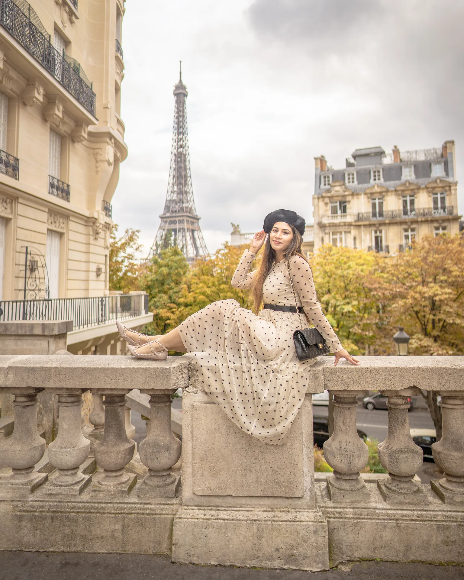 Woman in a polka-dot dress and beret posing on a Parisian stone balustrade with the Eiffel Tower and autumn trees behind her.