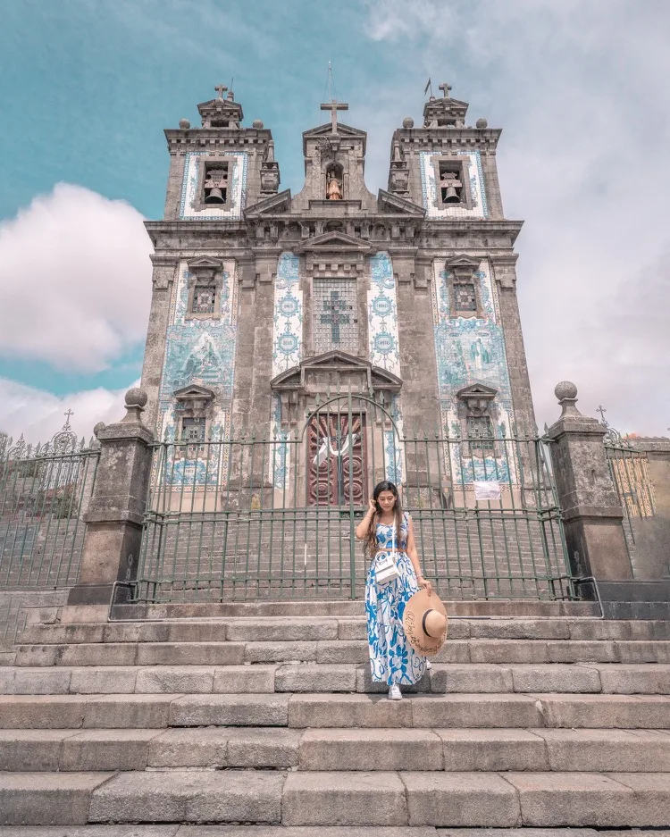 Young woman in a blue patterned dress holding a wide-brimmed hat as she descends stone steps in front of a tiled baroque church facade.