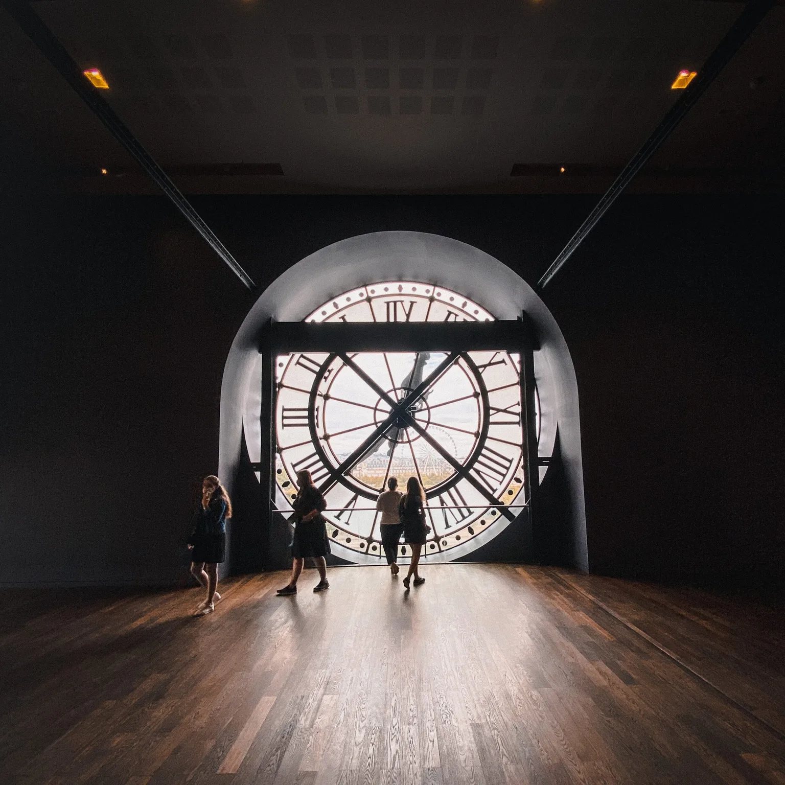 Visitors silhouetted against the giant ornate clock face window overlooking a cityscape inside a dim museum gallery.