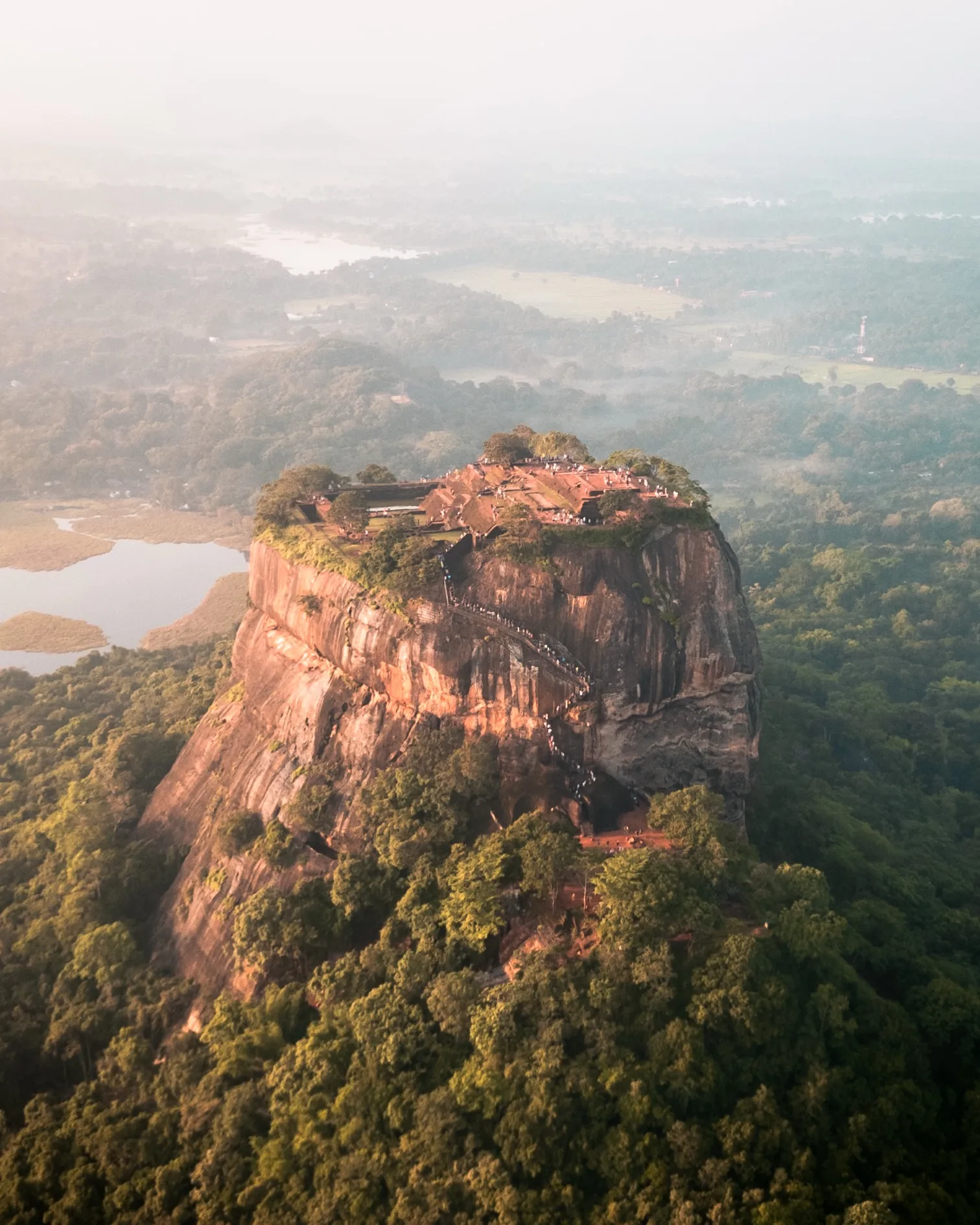 Ancient fortress and gardens atop a massive lone rock rising above misty tropical forest and nearby lakes at sunrise.