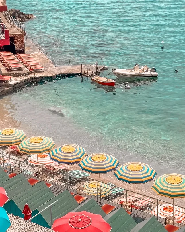 Colourful striped beach umbrellas and loungers line a rocky Mediterranean shore with two small boats floating in clear turquoise water.