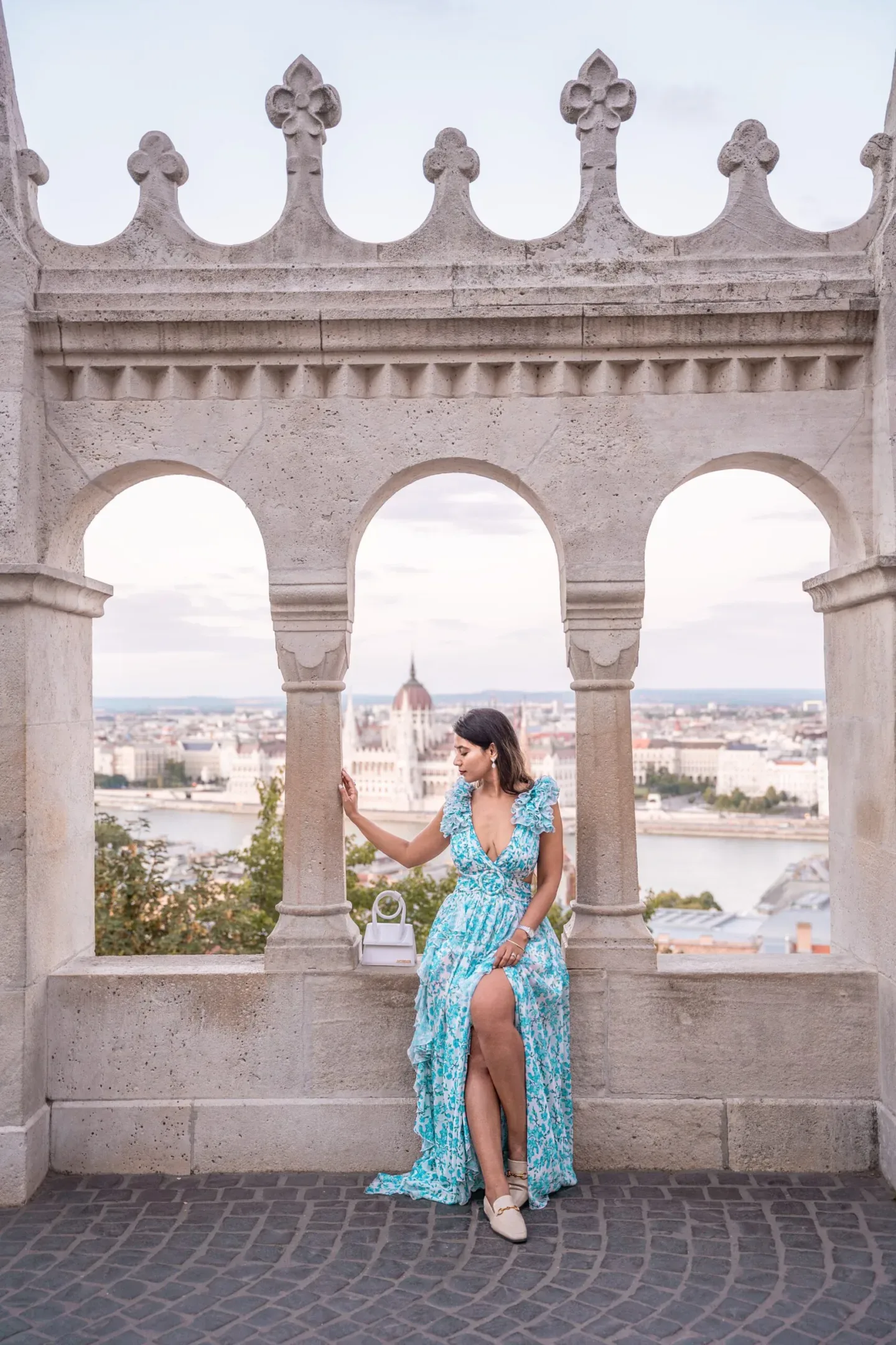 Woman in a flowing blue floral dress seated on a stone balustrade, framed by three arches overlooking the river and city skyline.