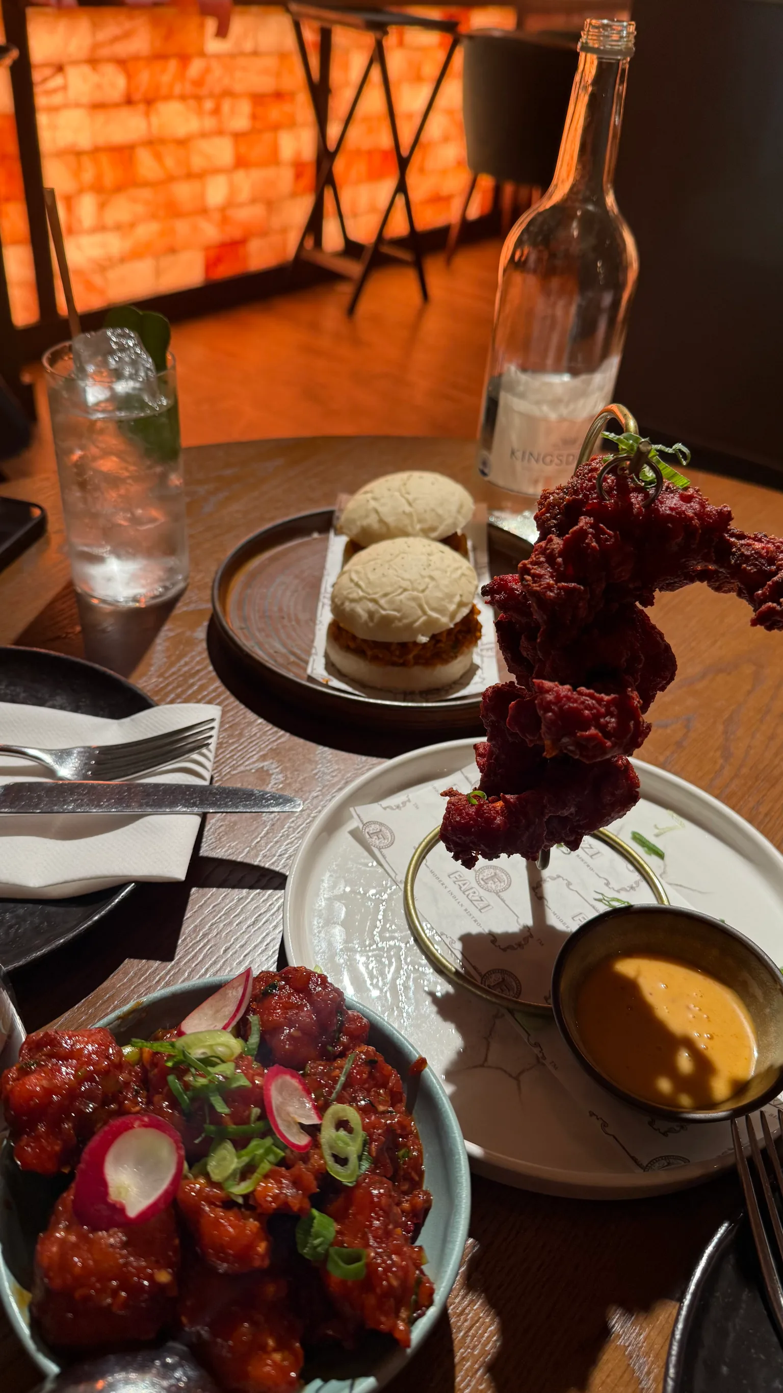 Assorted spicy fried chicken dishes and dipping sauce on a wooden table with two mini buns and a glass bottle in a warm-lit restaurant.
