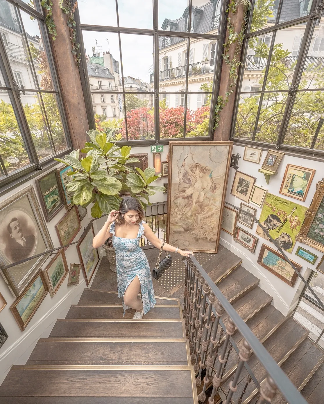 Woman in a floral dress ascending a sunlit, art-filled staircase with large windows overlooking Parisian rooftops.