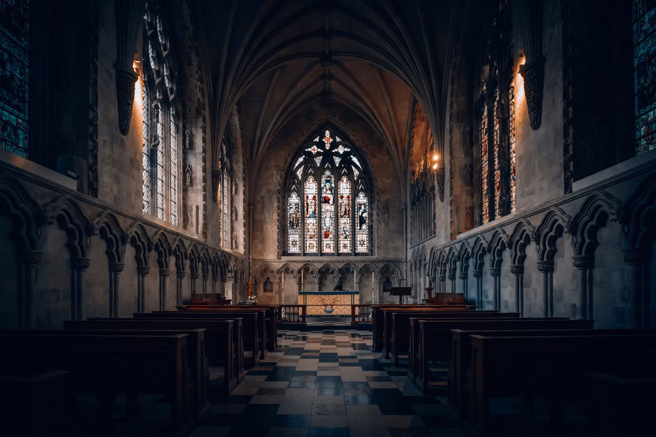 Dim Gothic church interior with stained glass windows, arched ceiling and pews leading to the altar