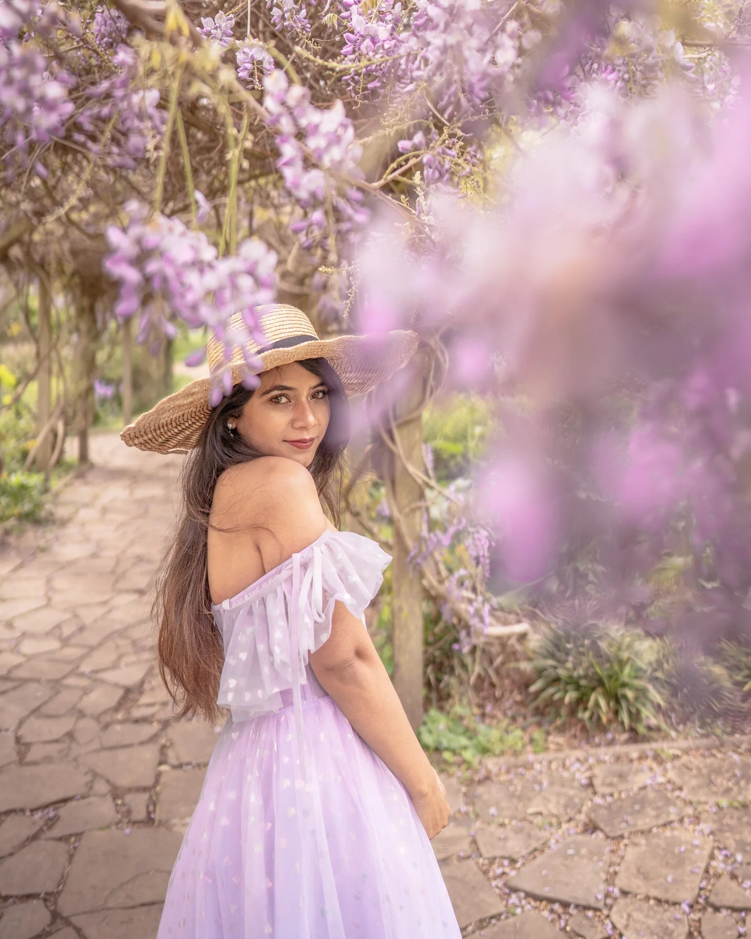 Woman in a straw hat and flowing lilac dress looking back under a canopy of wisteria along a stone garden path.