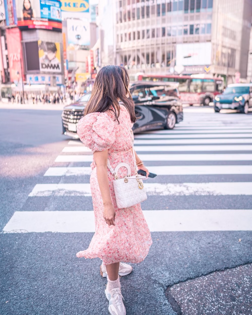 A woman in a pink floral dress crosses a busy city street at a large urban intersection