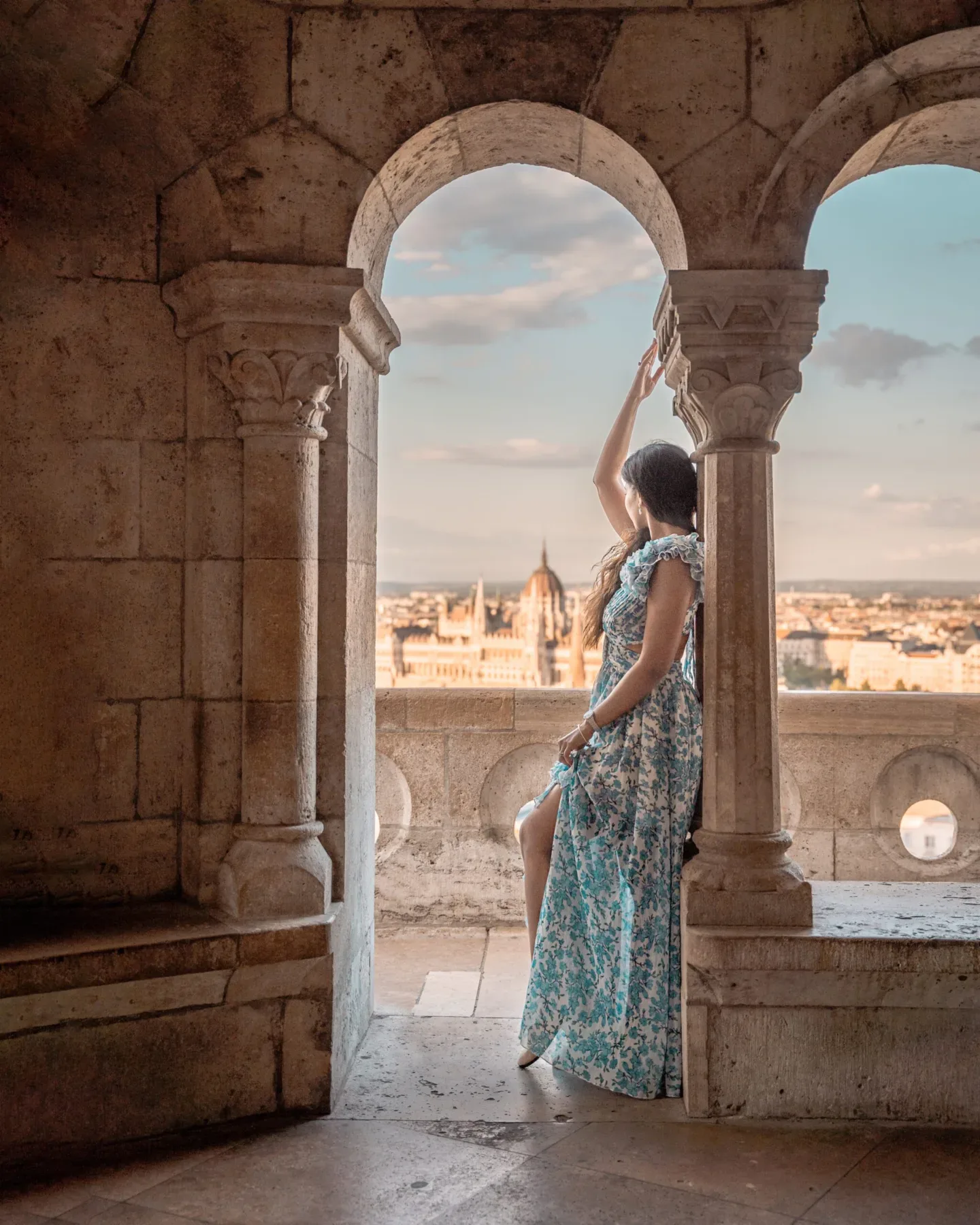 Woman in a blue floral dress standing in a stone archway overlooking a sunlit cityscape with a domed parliament building in the distance.